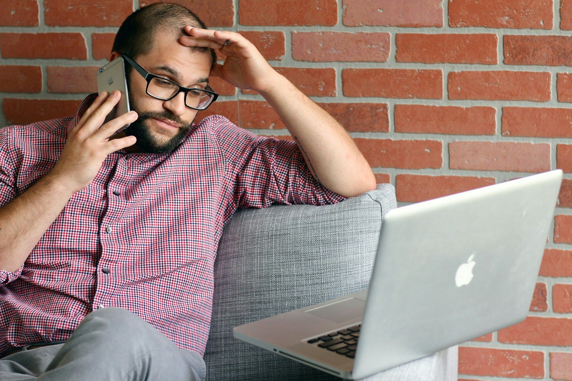 A man seated on a couch, using a laptop while holding a cell phone in his hand: via Unsplash