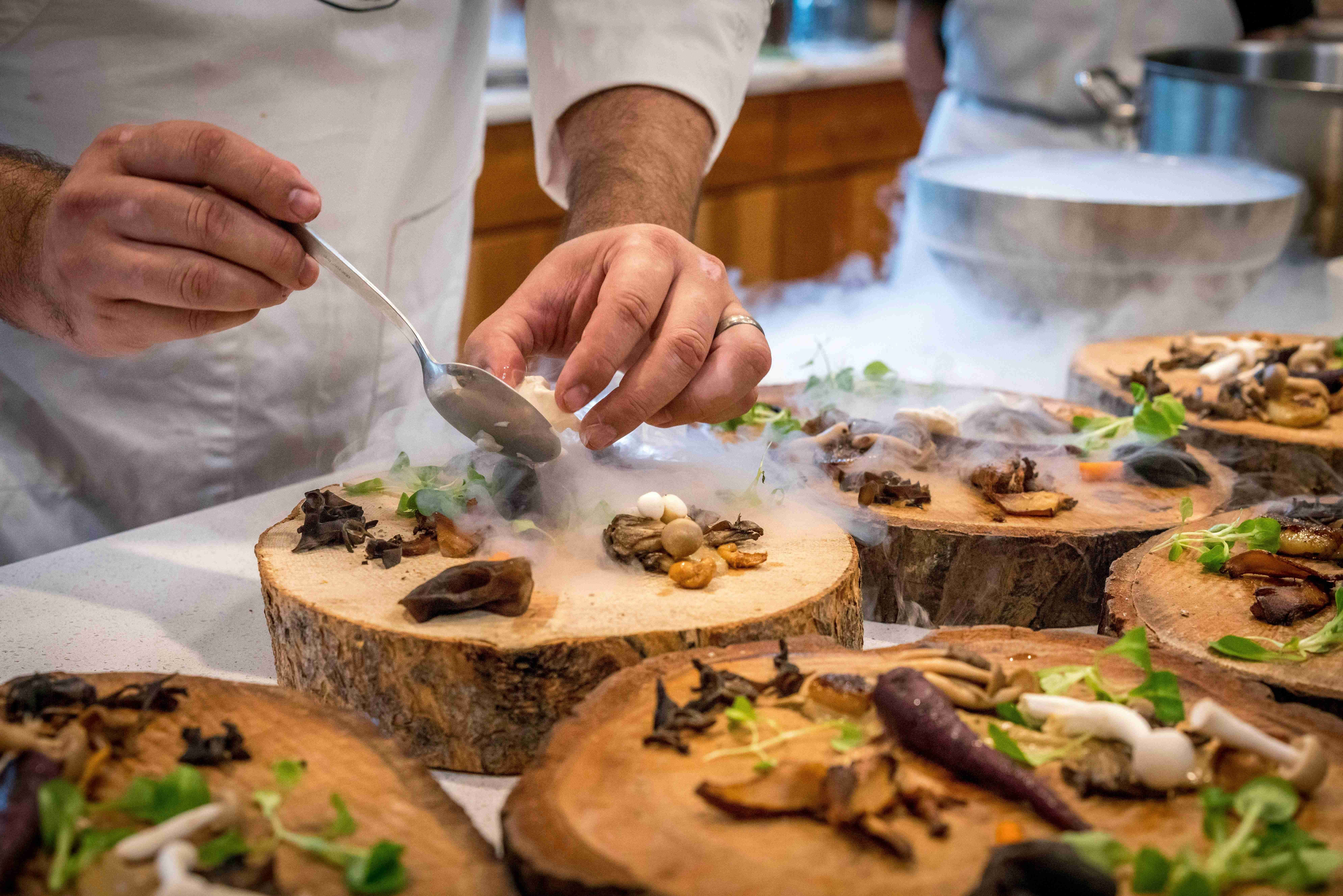 A chef meticulously plates a gourmet dish on a wooden slab, using a spoon to add final touches. (Image via Pexels)