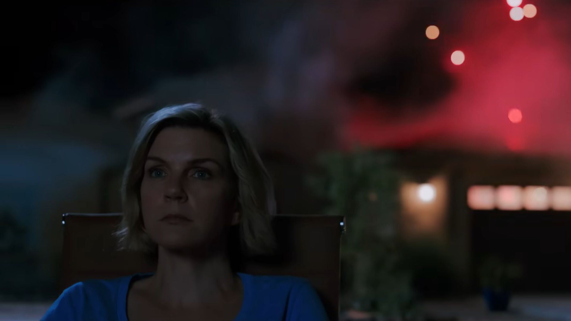 Carol is sitting with a blue t shirt on and flare-like smoke appears to be glowing in the dark background