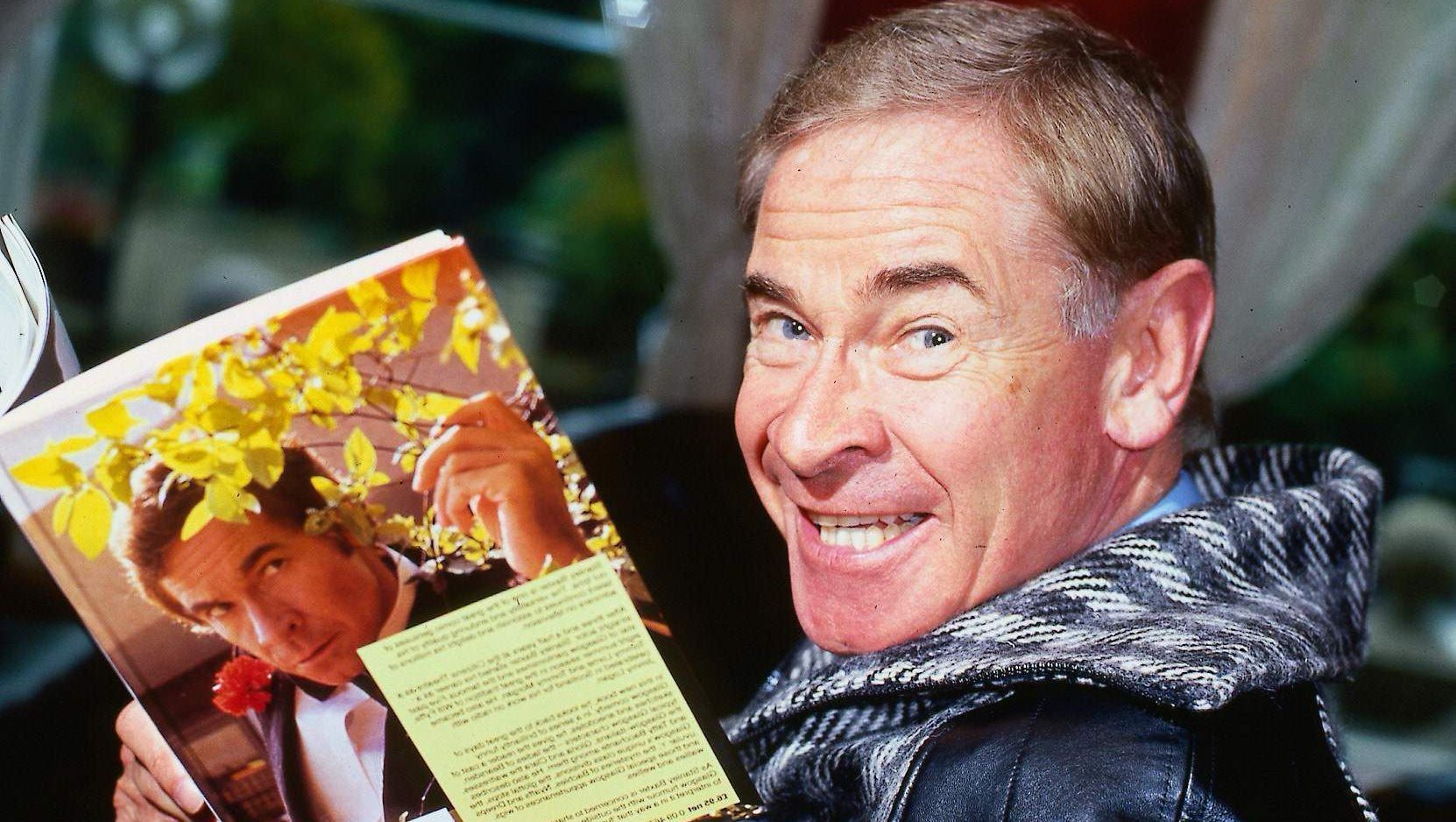 Stanley Baxter holding a book featuring his own portrait in September 1986. (Image via Getty/Mirrorpix)