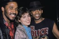 Kim Delaney, wearing a grey fur jacket over a turquoise blue outfit, and British actor Darnell Williams, who wears a 'The Police' t-shirt and a black fedora, United States, circa 1982 - Source: Getty